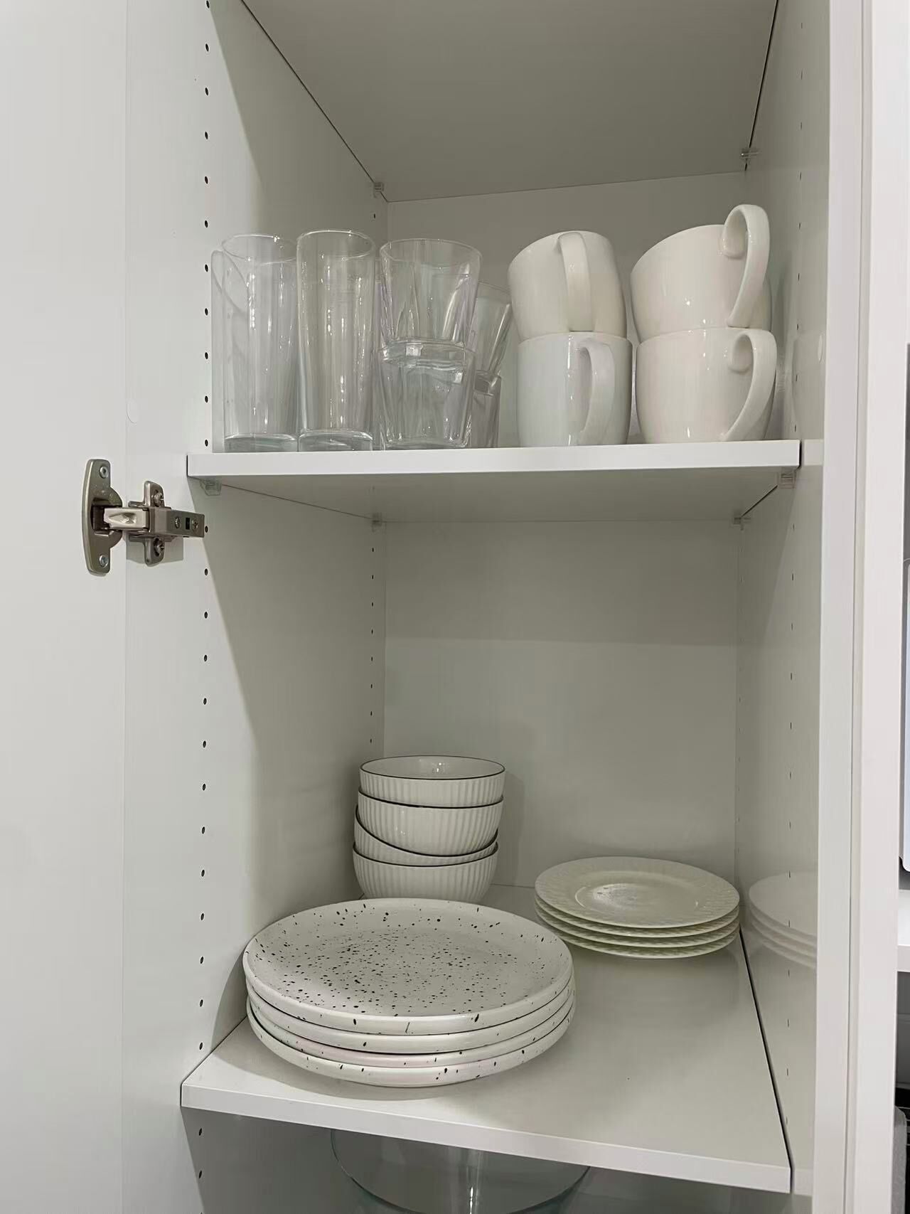 A white cabinet with shelves holding clear glassware, white cups, and stacked plates.