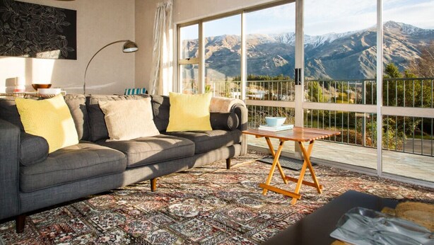 A living room with a gray sofa, yellow cushions, a patterned rug, a wooden coffee table, and a large window with a mountain view. - Kiwi Bach with Amazing Views (Wānaka)