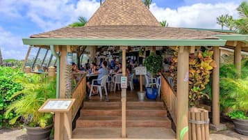 A covered outdoor seating area with a tiled roof, surrounded by potted plants and a sign indicating a waiting area.
