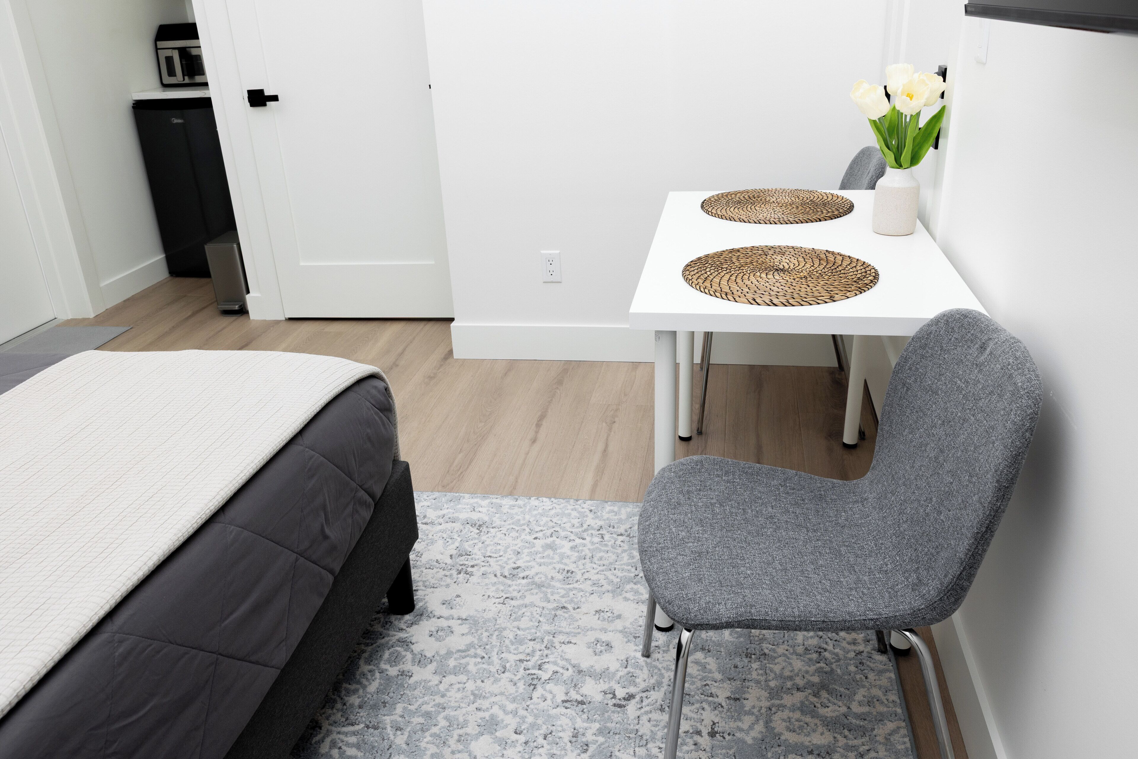 A bedroom with a bed, a gray chair, a small table with a vase of flowers, and a white wall.
