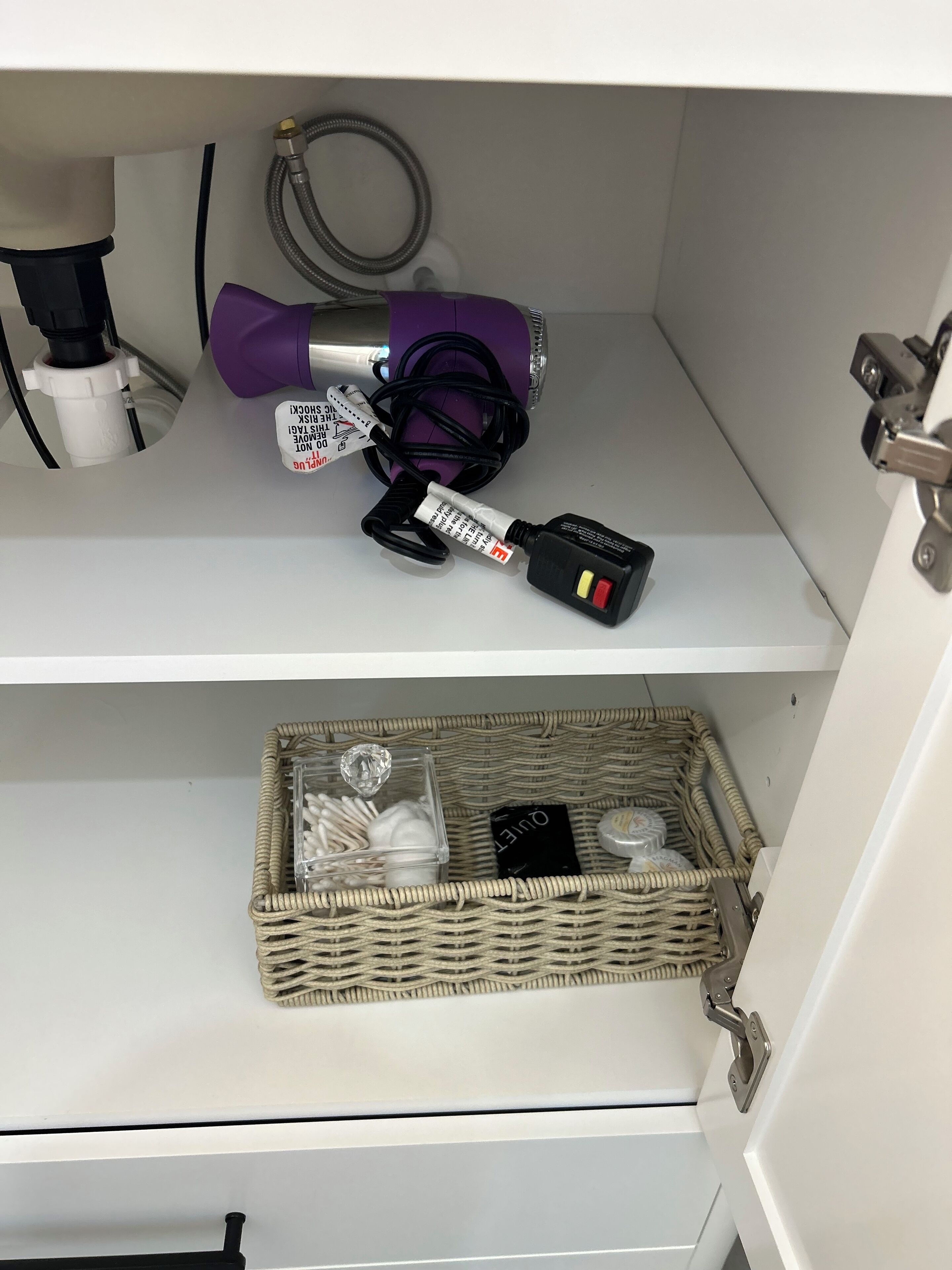 Un armario de almacenamiento con un dispositivo morado, un adaptador de corriente y una cesta de mimbre que contiene bolas de algodón y un pequeño objeto negro.
