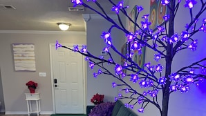 Un soggiorno con un divano color petrolio, un poggiapiedi verde e un elemento decorativo viola. Nell’angolo, un albero illuminato con luci viola.