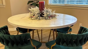 Un tavolo da pranzo rotondo con piano in marmo bianco, circondato da quattro sedie rivestite in verde con gambe dorate. Sul tavolo è posata una ghirlanda decorativa con pigne e bacche rosse, e una candela rossa alta è posta accanto.