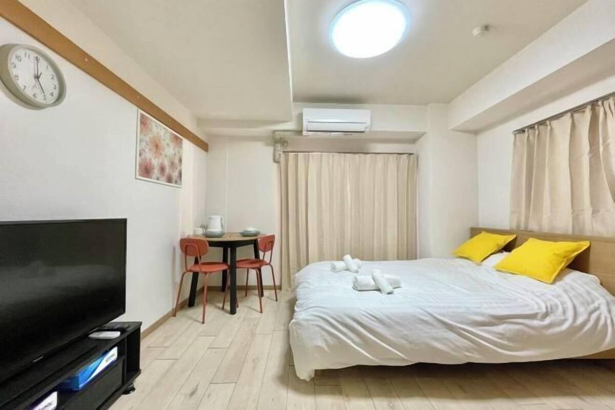 A hotel room with a bed, a TV, a desk with chairs, and an air conditioning unit.