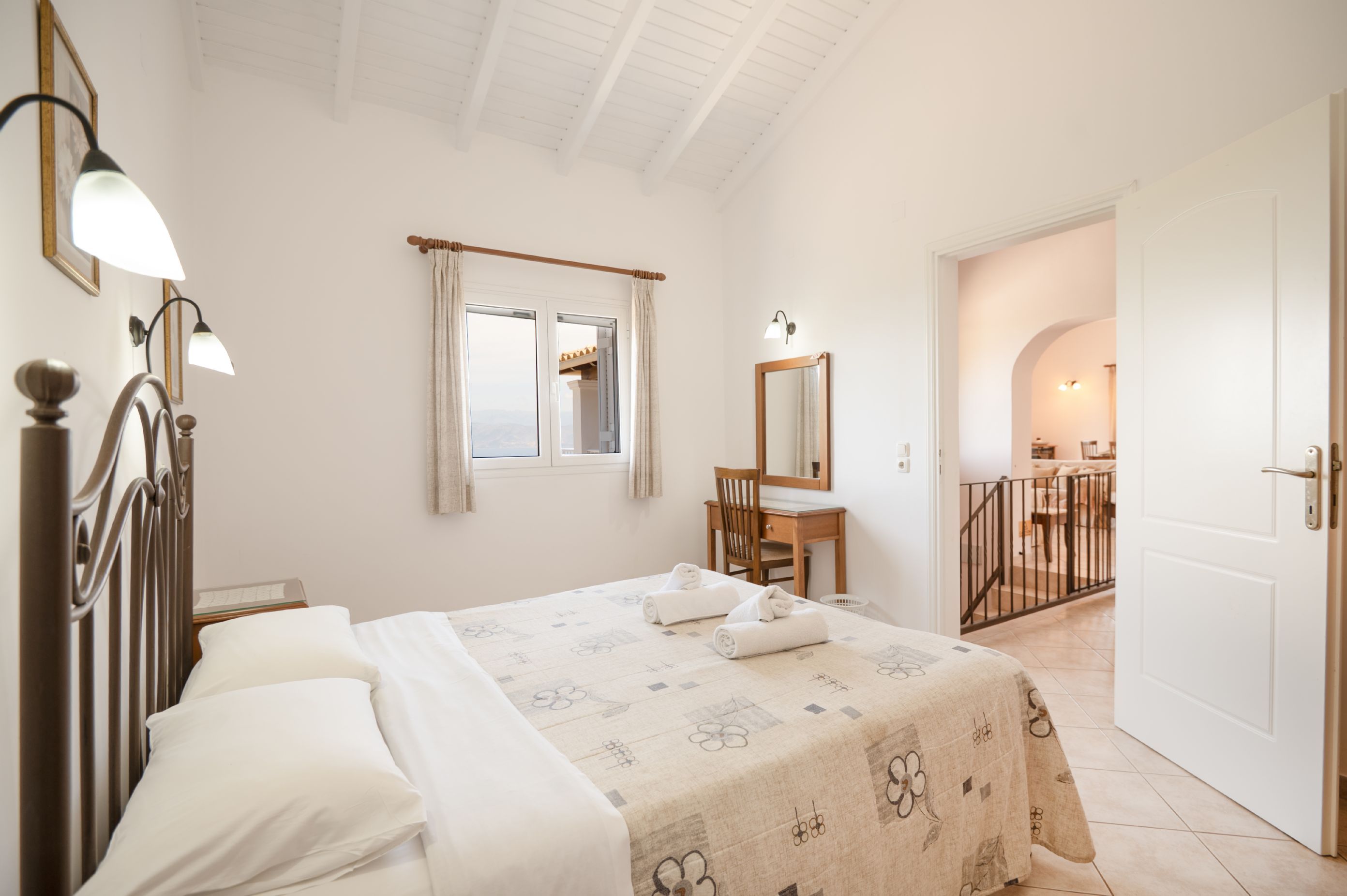 A hotel room with a bed, a desk, a chair, a window with curtains, a mirror, and a door leading to another room.