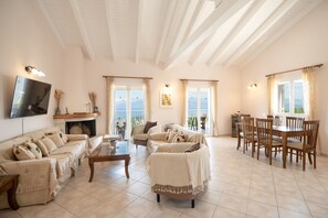 A spacious living room with a fireplace, a dining area with wooden furniture, and large windows offering a view of the outdoors. - Corfu Holiday Villa with Pool, Sea Views, Family Friendly (Kassiopi)
