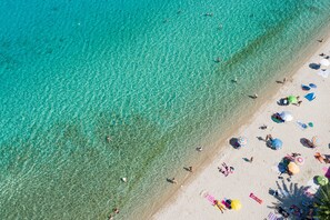 Ovenudblik over en strand med krystalklart turkis vand, folk der solbader, og farverige parasoller.