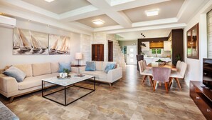 A spacious living area with a sofa, coffee table, and dining table set. There is a kitchen in the background and a large window providing natural light. - Sunny Exclusive Ena – Villa with Private Pool & Sea Views (Hanioti)
