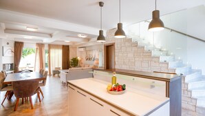 A modern kitchen with a central island, pendant lighting, and a staircase with glass railings. - Sunny Exclusive Ena – Villa with Private Pool & Sea Views (Hanioti)
