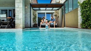 A modern outdoor pool area with a stone wall, a covered seating area, and a view of the surrounding landscape. - Sunny Exclusive Ena – Villa with Private Pool & Sea Views (Hanioti)