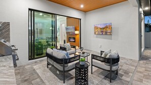 Eine moderne Außenpatio mit einer Couch, Sesseln und einem kleinen Tisch, mit einer Glastür zu einem Innenbereich mit Fernseher und Gemälde.