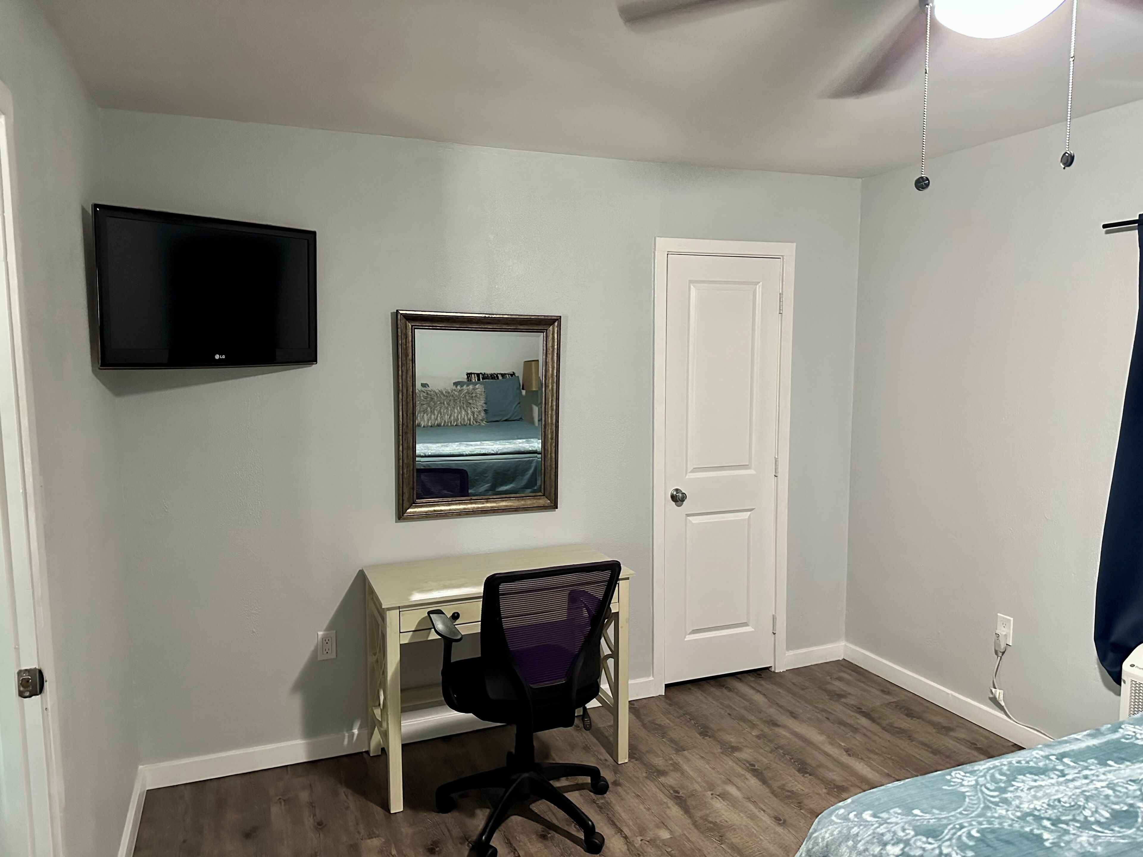 A bedroom with a bed, a desk, a chair, a TV, a door, and a mirror.