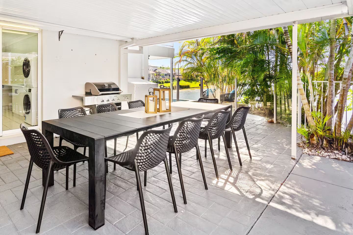 An outdoor patio with a dining table and chairs, a grill, and a view of a garden with palm trees.