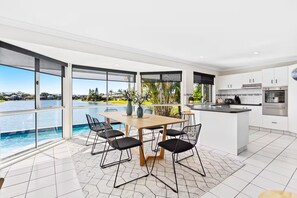 A modern kitchen with large windows overlooking a pool, a dining area with a table and chairs, and a view of a residential area. - Stunning Waterfront Home - 3 Large Ensuites! (Broadbeach Waters)
