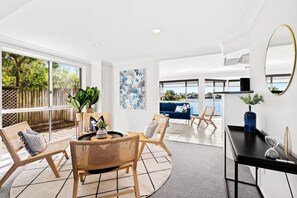 A modern living room with a large window, a blue sofa, wicker chairs, and a black coffee table. - Stunning Waterfront Home - 3 Large Ensuites! (Broadbeach Waters)