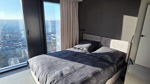 Ett modernt sovrum med en stor säng, en skjutdörr i glas som leder ut till en balkong och utsikt över staden.