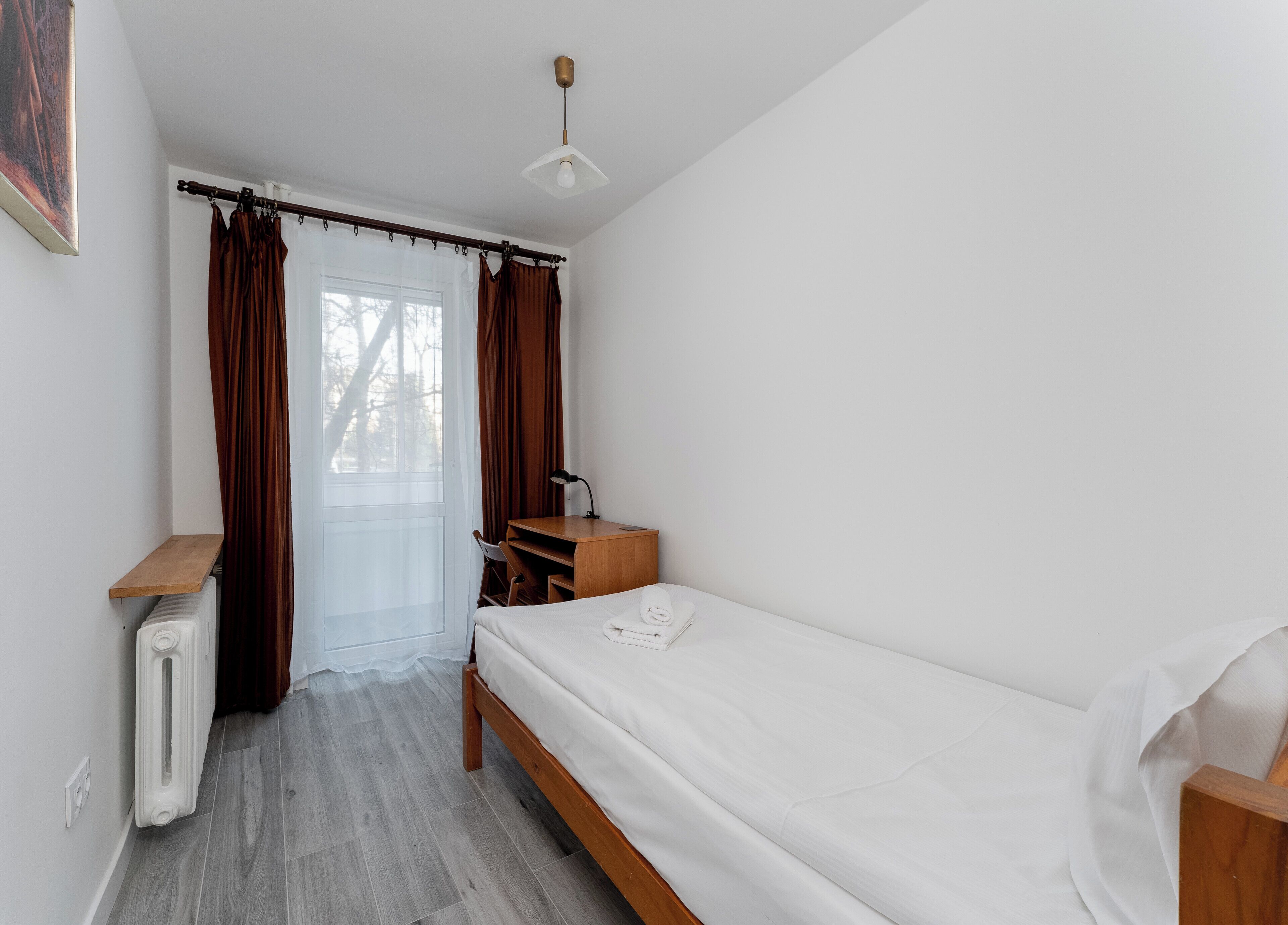 A small, tidy room with a single bed, a wooden desk, a chair, a radiator, and a window with curtains.