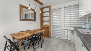 A modern kitchen with a wooden dining table, black chairs, a built-in shelf unit, and a window with blinds. - Bronowice Spacious Apartment (Kraków)