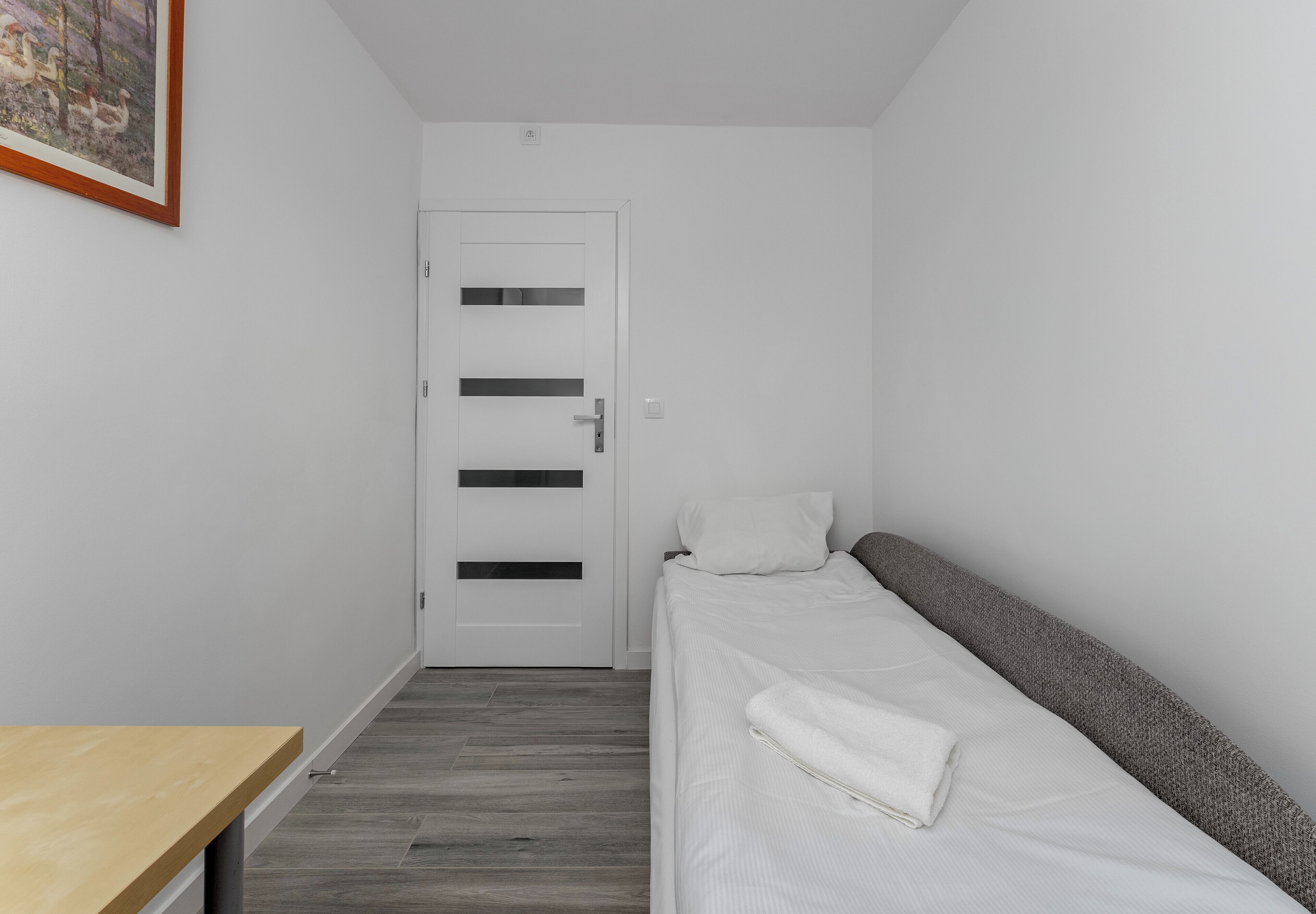 Ein kleiner, moderner Raum mit einem Bett, einem Holztisch und einer Tür mit Lamellen.