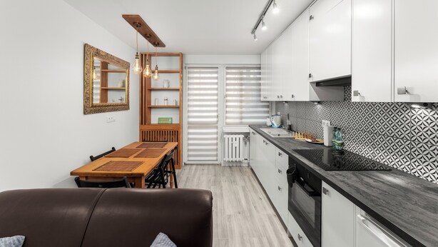 A modern kitchen with white cabinets, a black countertop, and a patterned backsplash. There is a dining area with a wooden table and chairs, and a brown sofa. - Bronowice Spacious Apartment (Kraków)