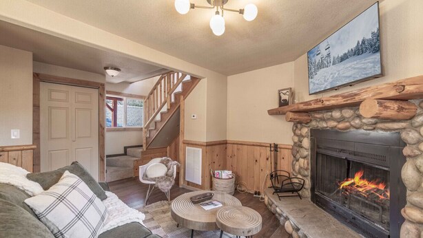 A cozy living room with a fireplace, a staircase, and a large window. - Bear Den|Jacuzzi, AC, Steps from trails, Dogs OK! (Big Bear)