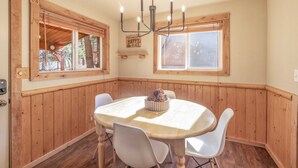 A cozy dining area with a wooden table and chairs, a chandelier, and a window with a view of trees. - Bear Den|Jacuzzi, AC, Steps from trails, Dogs OK! (Big Bear)