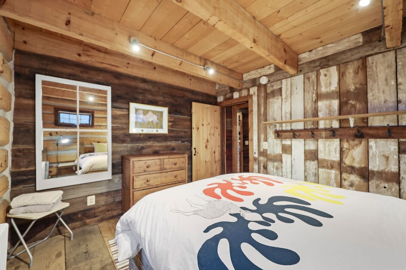Une chambre chaleureuse dans un chalet en rondins, avec un plafond en bois, un lit recouvert d’une couverture colorée, une commode et un miroir qui reflète la pièce.