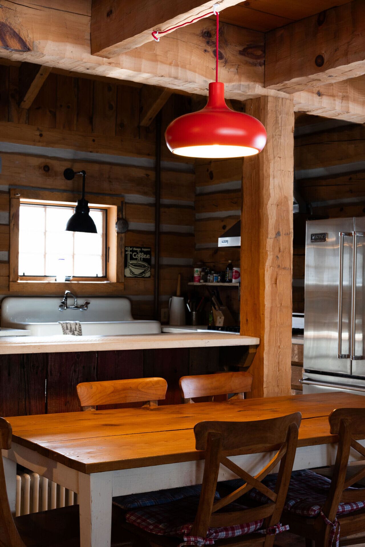 L’intérieur d’un chalet rustique est doté d’une table à manger en bois, de chaises, d’une suspension rouge, d’une fenêtre avec une lampe noire et d’un réfrigérateur en acier inoxydable.