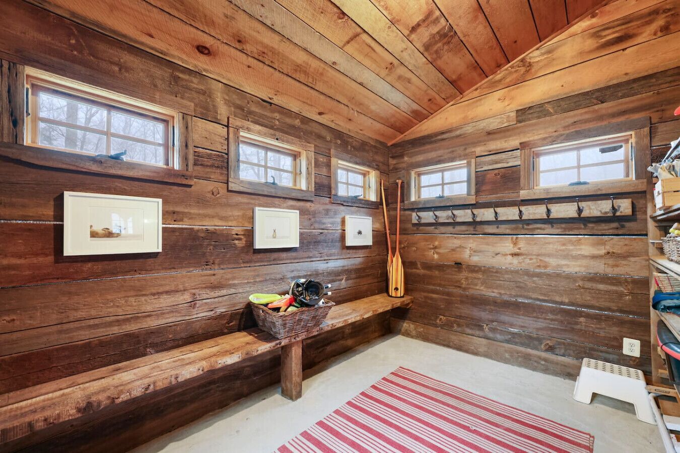 L’intérieur d’une cabane avec des murs en bois, de nombreuses fenêtres, un banc, un panier contenant divers objets et un tapis à rayures rouges.