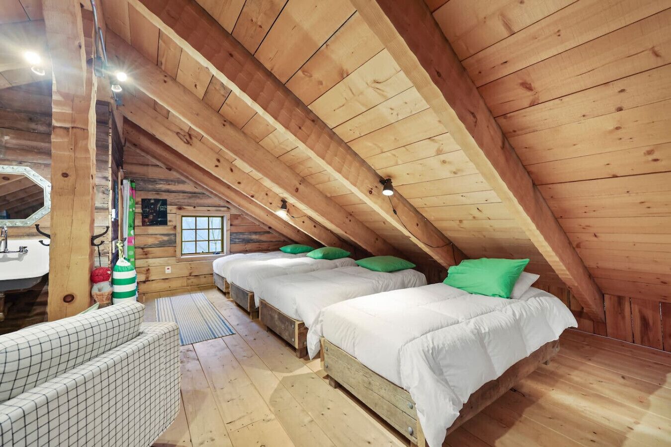 Une chambre mansardée chaleureuse avec des poutres en bois, deux lits avec des oreillers verts, une petite fenêtre et un lavabo.