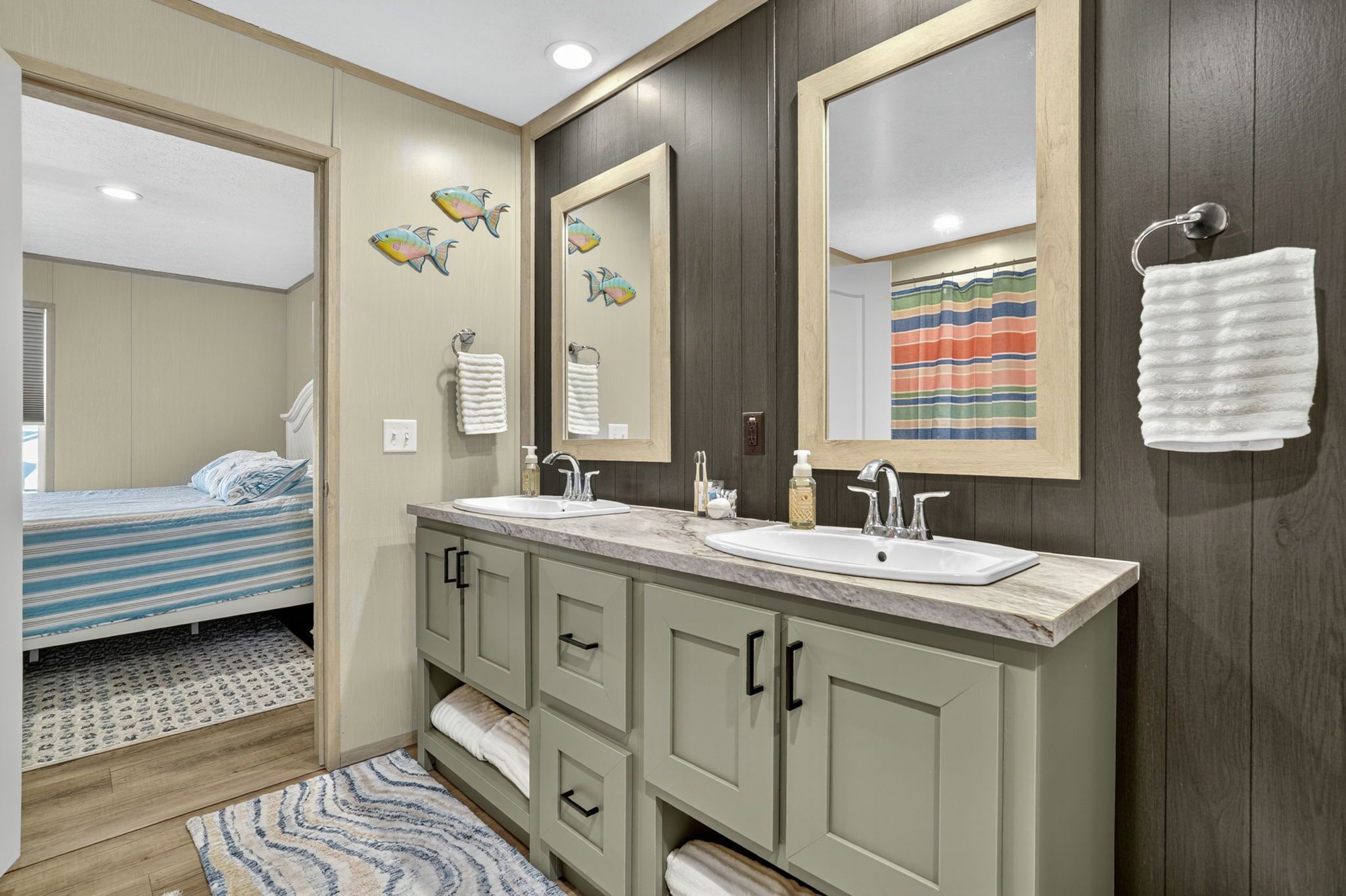 A bathroom with two sinks, a large mirror, and a bed in the adjacent room.