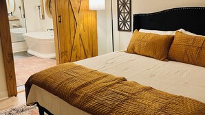 Un dormitorio con una cama grande, una puerta de madera, un espejo y un baño visible a través de una puerta abierta.