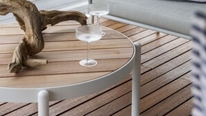 Una terrassa de fusta amb una taula rodona, dos vasos i un objecte decoratiu. Un sofà gris amb coixins estampats es troba al fons, amb vista a un paisatge verd.