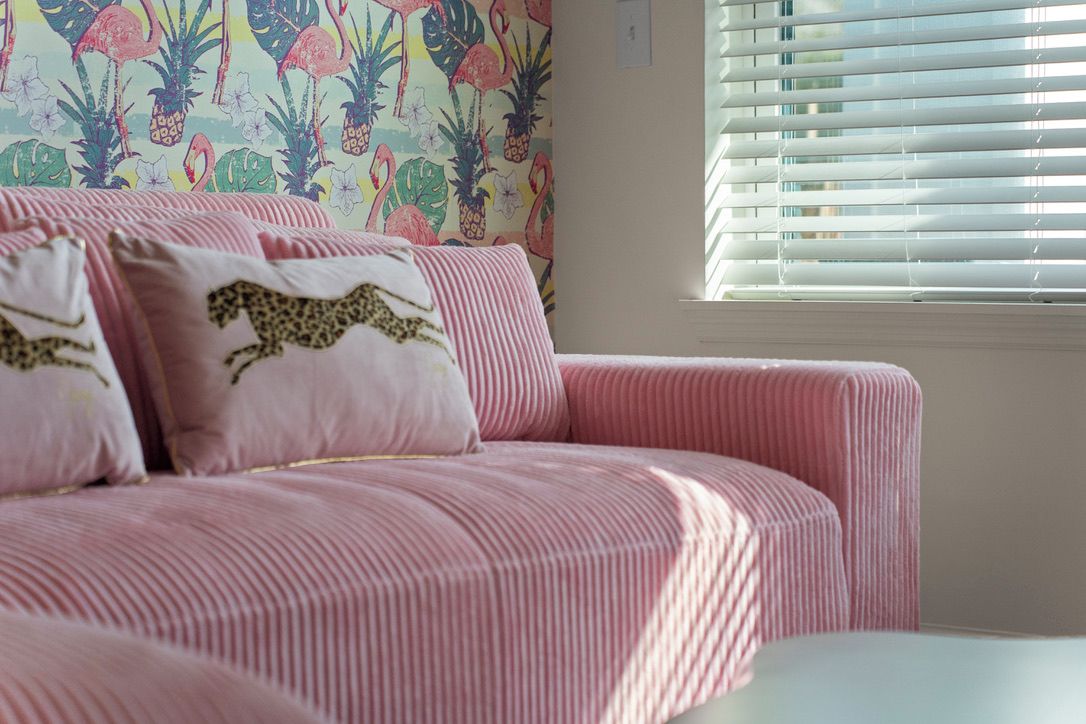 Un canapé rose recouvert de tissu, agrémenté de coussins à motifs léopard, placé devant un mur orné d’un papier peint aux motifs tropicaux.