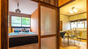A bedroom with a bed, bedside table, and a view through a sliding door to a kitchen with a dining table and chairs. - Showa House – Spacious Retro Stay near Haneda (Ota City)