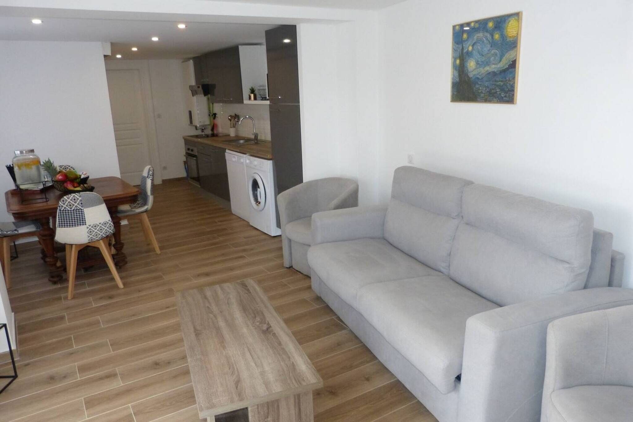 A modern living room with a grey sofa, a wooden coffee table, and a dining area with a wooden table and chairs. A washing machine is visible in the background.
