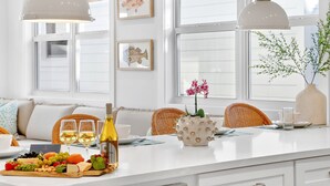 Een goed verlicht eetgedeelte met een witte tafel gedekt voor een maaltijd, met verschillende soorten fruit, kaas en wijn, plus twee hangende hanglampen en een wanddecoratie van viskunst.