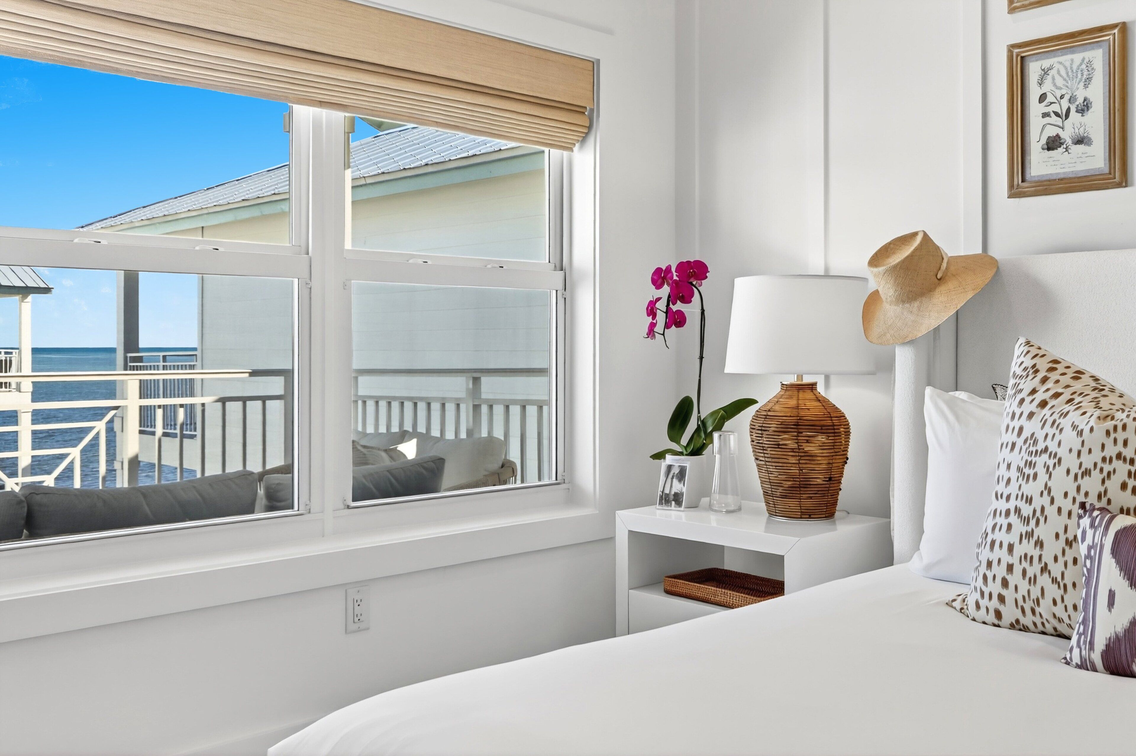 A hotel room with a large window overlooking a balcony, a bed with patterned pillows, a bedside table with a lamp and decorative items, and a framed botanical print on the wall.
