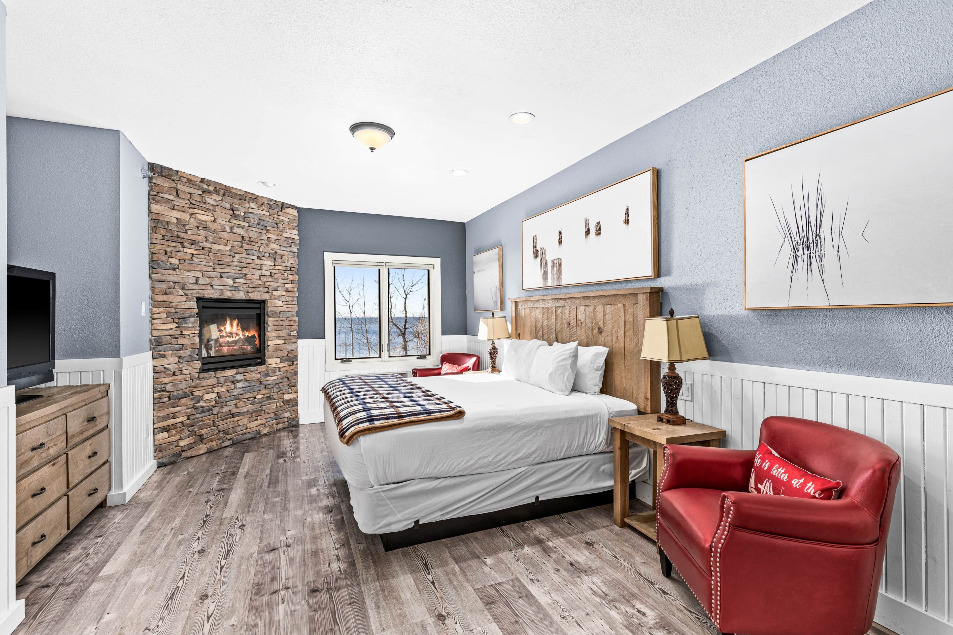 A bedroom with a stone fireplace, a bed, a red armchair, and artwork on the walls.