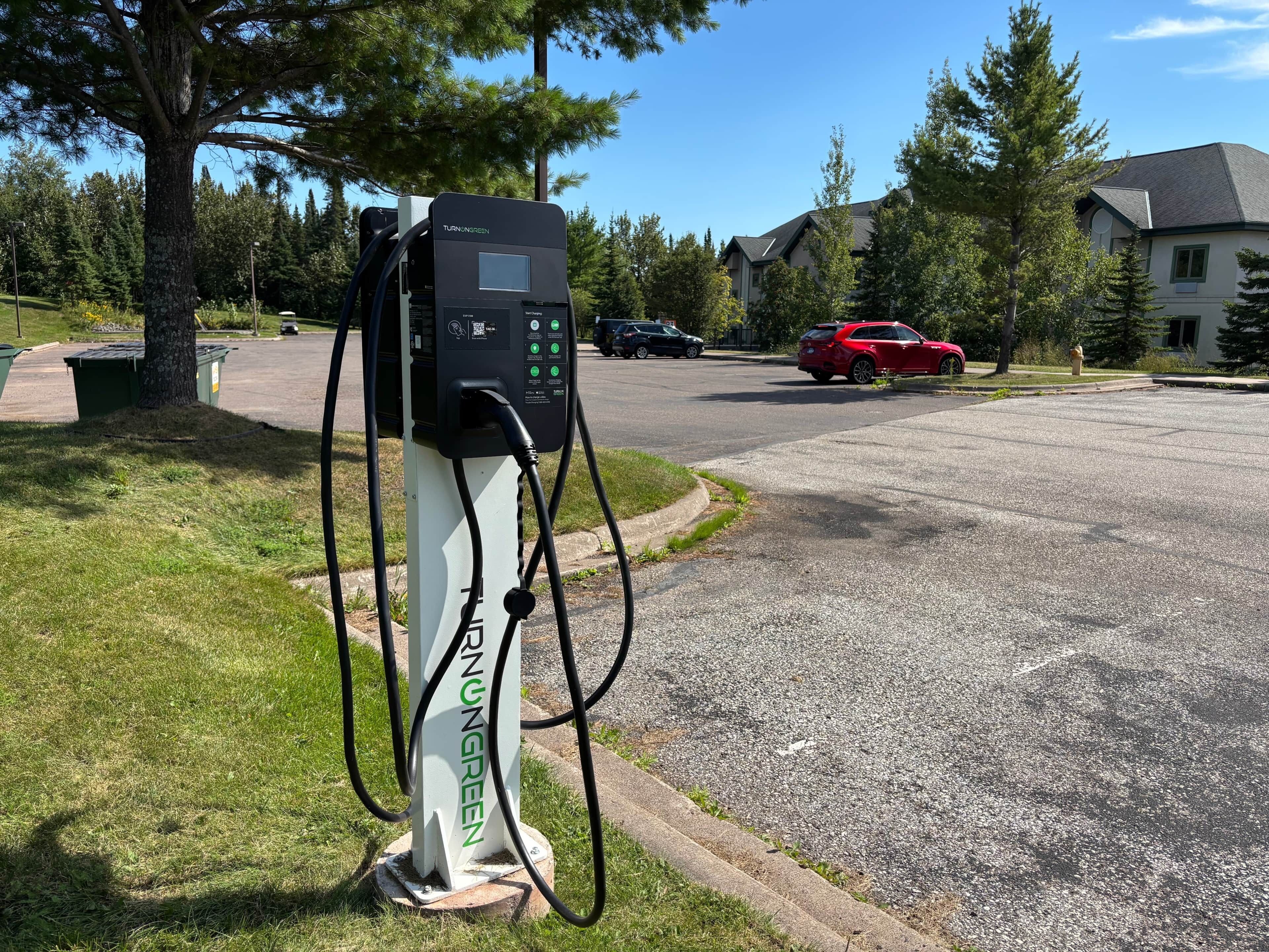 A charging station with a digital display, located in a residential area with houses and a red car parked nearby.