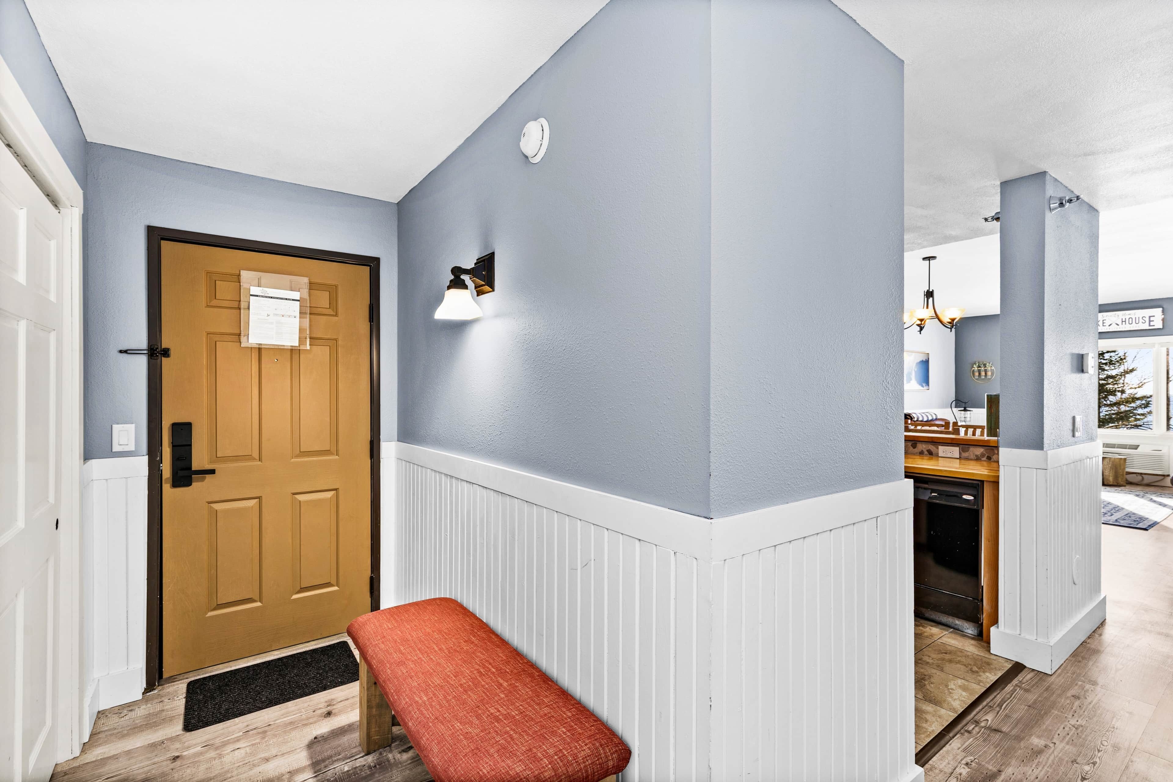 A hallway with a wooden door, a bench, a wall-mounted light fixture, and a kitchen area visible through an open door.