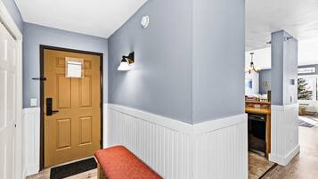 A hallway with a wooden door, a bench, a wall-mounted light fixture, and a kitchen area visible through an open door.