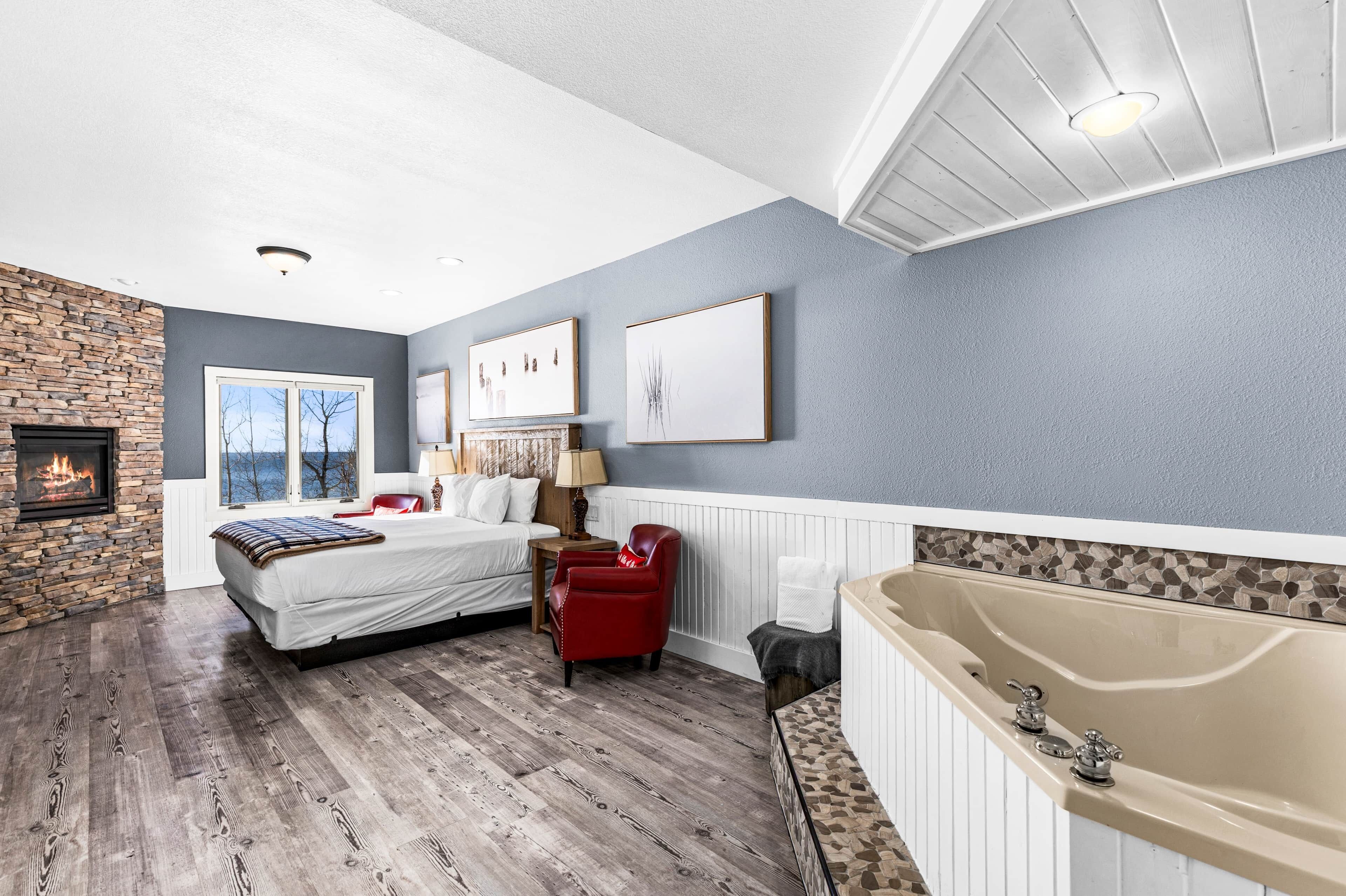 A bedroom with a stone fireplace, a large bed, a red chair, a bathtub, and framed artwork on the wall.