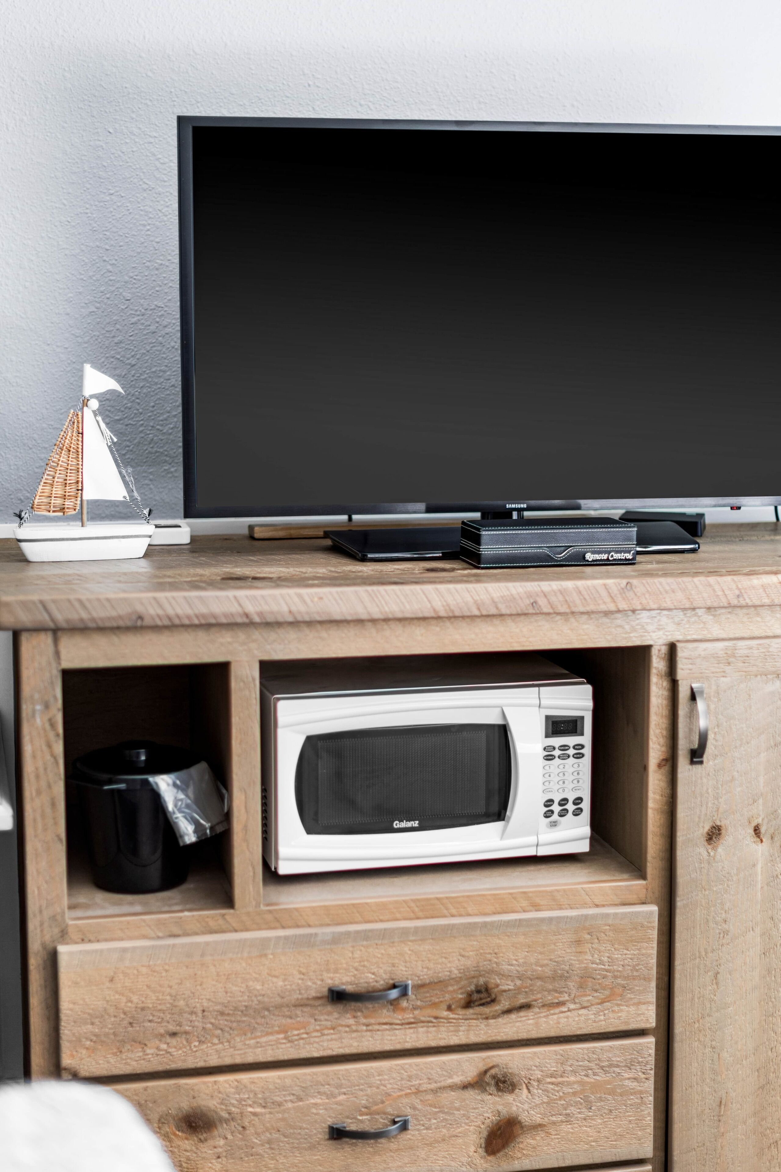 Un meuble en bois sur lequel sont posés un four à micro-ondes, une maquette de petit voilier et une télécommande Samsung.