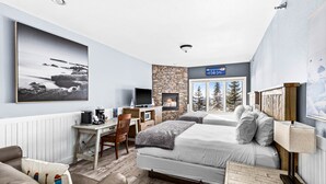 Une chambre d’hôtel moderne avec une cheminée en pierre, un grand lit, un bureau avec un ordinateur et une vue sur les arbres par la fenêtre.