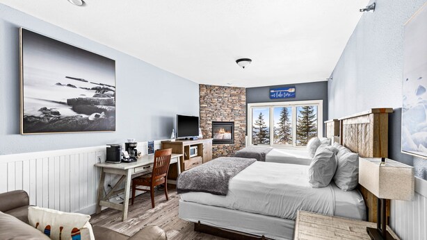 A modern hotel room with a stone fireplace, a large bed, a desk with a computer, and a view of trees through the window. - Whispering Pines Lake Views | 2 Queen Bed Studio (Two Harbors)