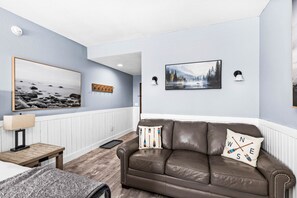 A living room with a brown leather sofa, a wooden coffee table, a lamp, and wall-mounted artwork. - Whispering Pines Lake Views | 2 Queen Bed Studio (Two Harbors)