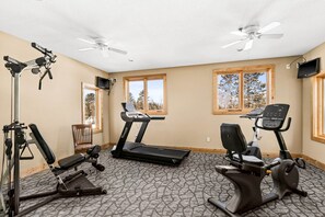 Un gimnasio con cintas de correr, elíptica y bicicleta estática. Hay ventiladores de techo, un televisor y ventanas amplias con vistas a árboles.