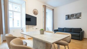A modern living room with a dining table, chairs, a sofa, a flat-screen TV, and a potted plant. - Cozy 1BR apartment near Opera - 24h self check-in (Wien)