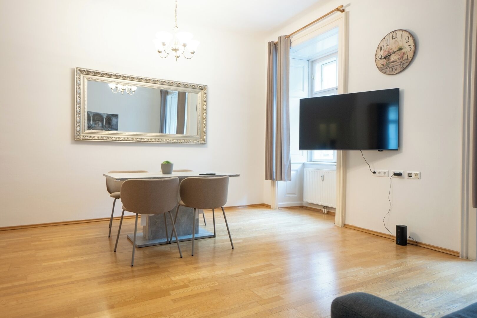 A modern living room with a dining table, chairs, a flat-screen TV, a large mirror, and a chandelier.